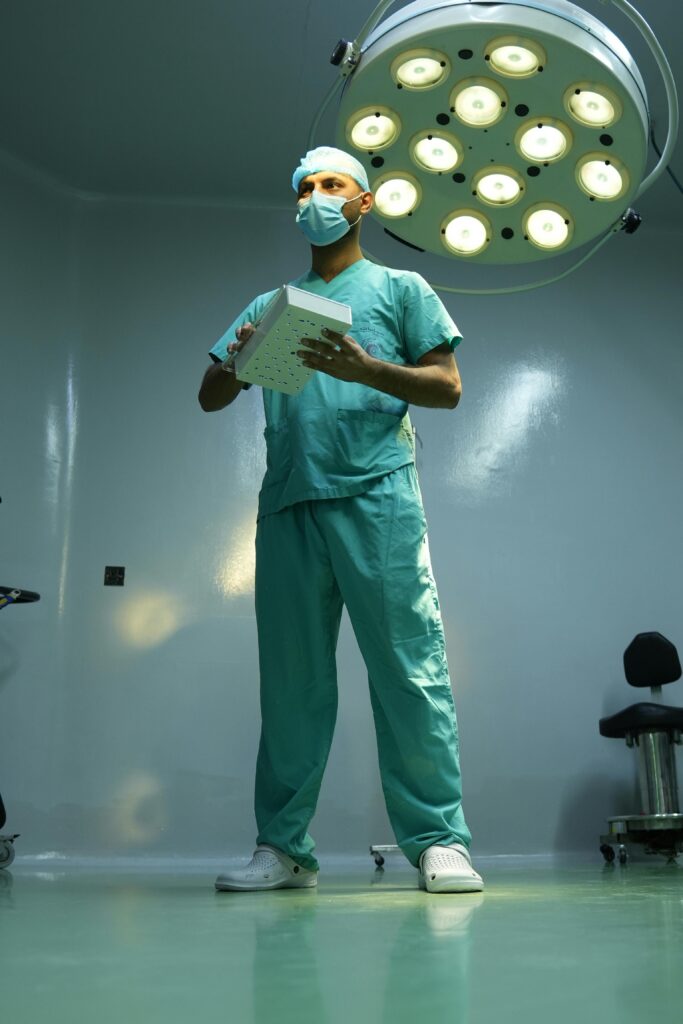 A surgeon in scrubs and mask stands ready in a well-lit operating room.